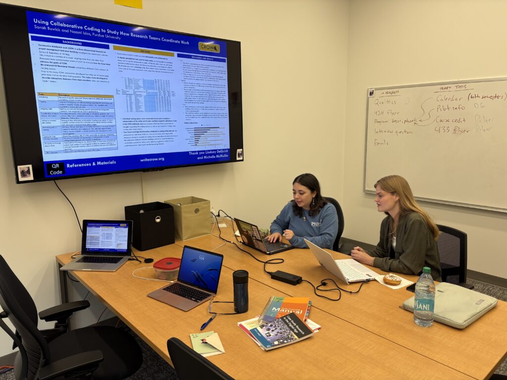 Two Purdue Crowbirds, Sarah Buwick and Naomi Islas, are sitting at a large table filled covered with other laptops and books, located underneath a nice flatscreen TV. They are working on their computers, and exchanging information. 