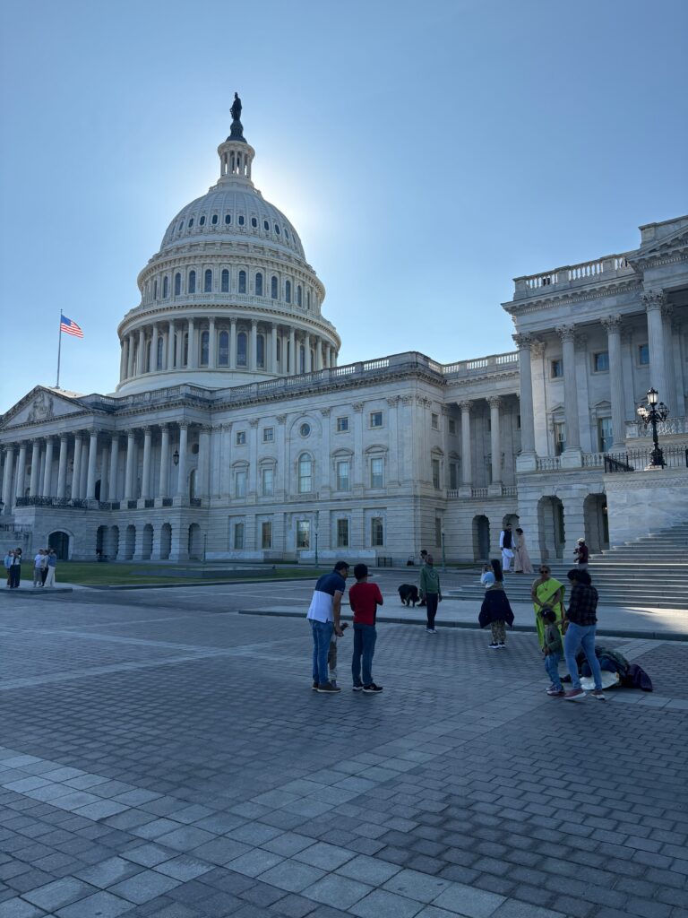 A bright blue sky, with the sun being blocked by the grand U.S. Capitol Building. It's a structure comprised of many pillars and rectangular windows, with the American Flag mounted on one of the rooftops. There is also a multi-colored brickyard beyond the steps of the building where groups of people chat. 