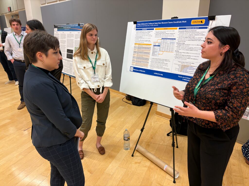 Two Purdue researchers, Sarah Buwick and Naomi Islas, stand in front of their poster. Naomi is presenting to an audience member, who is listening attentively, while Sarah, also listening, stands with her hands clasped.