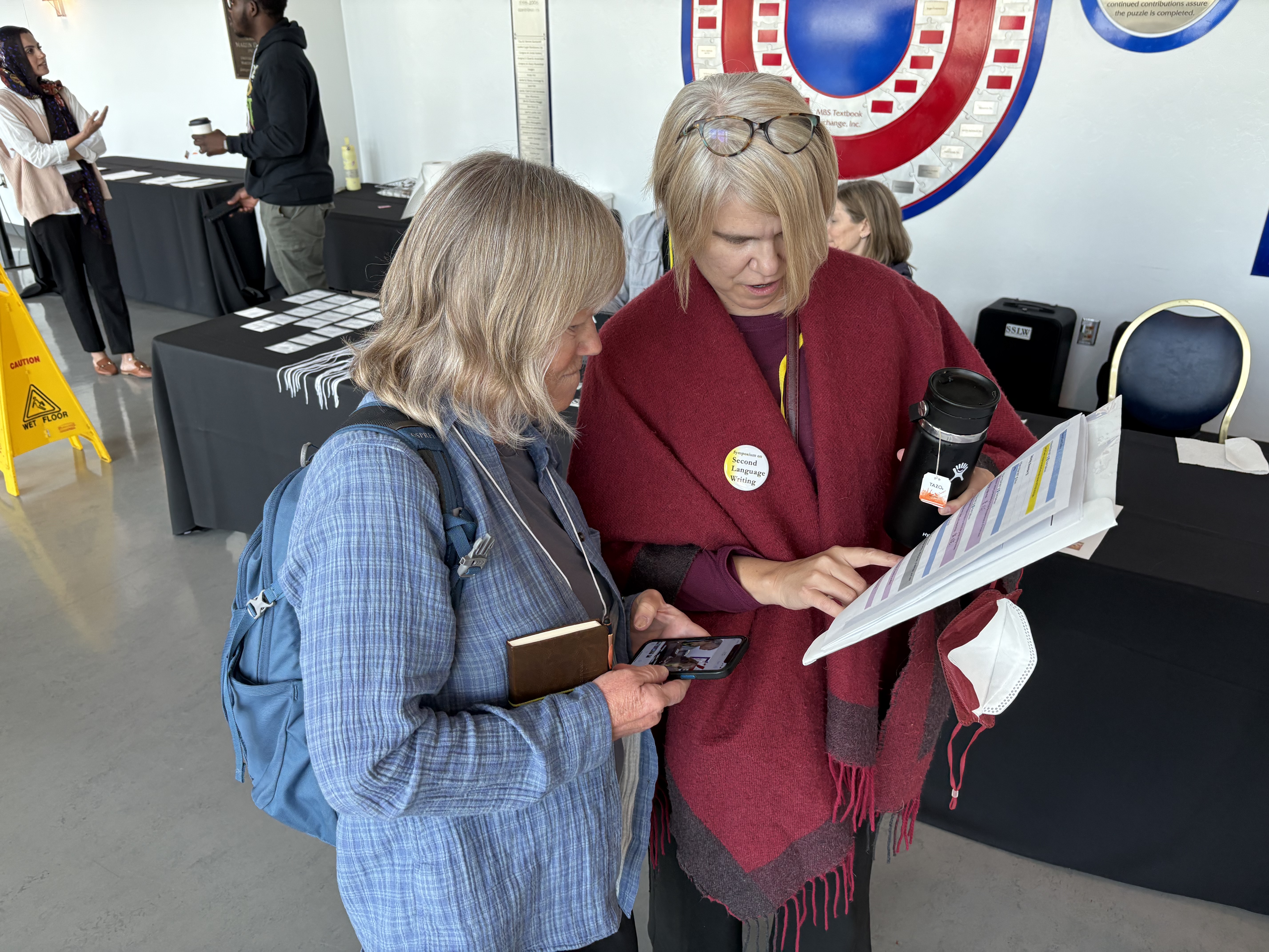 Dr. Shelley Staples is wearing a red shawl and is looking down while pointing to a document that she is holding. Dr. Randi Reppen stands closely next to her, in a blue long sleeve shirt, while looking at the document.