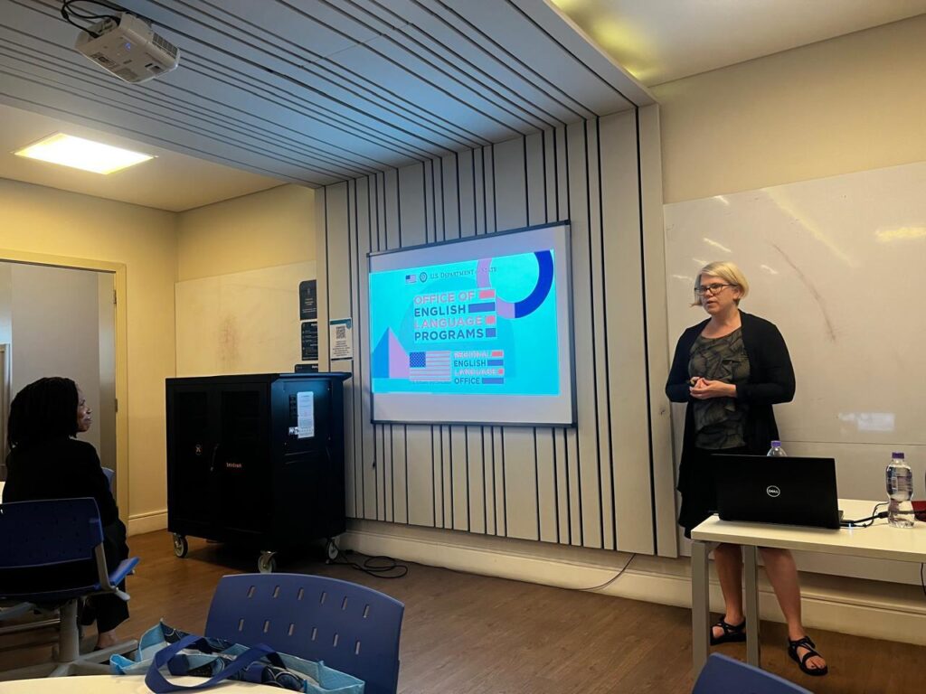 Dr. Shelley Staples stands in a classroom facing a few visible tables and people at PUC-SP. She is  in front of a whiteboard and projector that displays "Office of English Language Programs." Dr. Staples is wearing a black cardigan and sandals while her hands are clasped across her abdomen.
