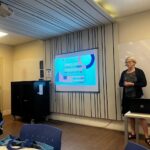 Dr. Shelley Staples stands in a classroom facing a few visible tables and people at PUC-SP. She is in front of a whiteboard and projector that displays "Office of English Language Programs." Dr. Staples is wearing a black cardigan and sandals while her hands are clasped across her abdomen.