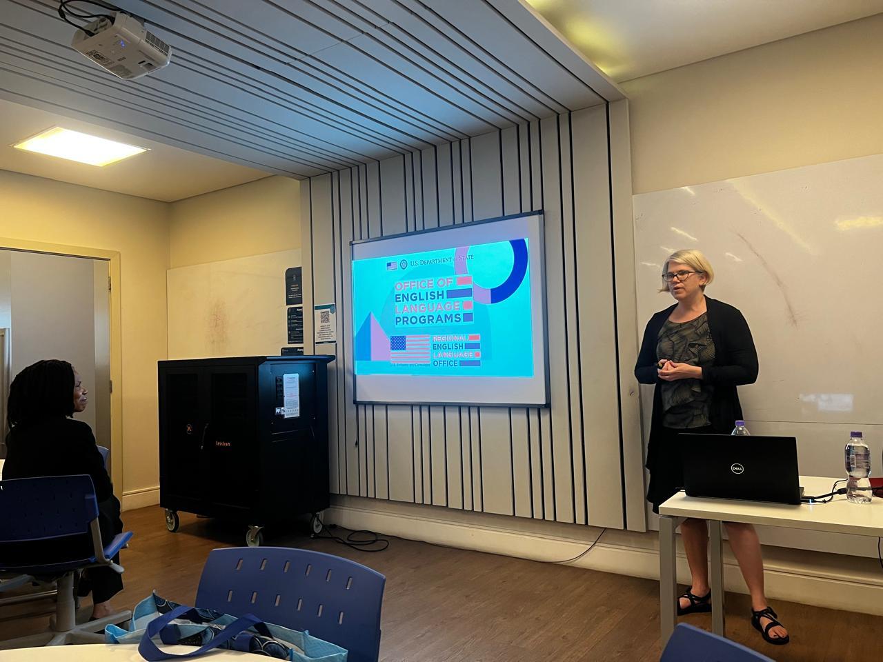 Dr. Shelley Staples stands in a classroom facing a few visible tables and people at PUC-SP. She is in front of a whiteboard and projector that displays "Office of English Language Programs." Dr. Staples is wearing a black cardigan and sandals while her hands are clasped across her abdomen.